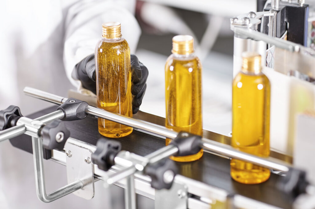 Pharmaceutical and Nutraceutical. Three clear bottles filled with yellow liquid are being lined up on a conveyor belt in an automated bottling process. A person wearing a glove is adjusting one of the bottles. The machinery is metallic and industrial.