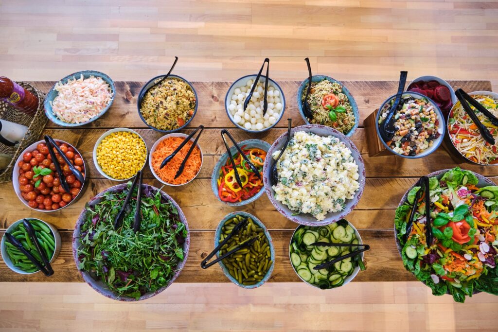 A wooden table brimming with delectable food, showcasing an array of salads and toppings: arugula, cucumber slices, tomatoes, corn, pasta salad, potato salad—all beautifully presented in bowls with serving utensils.
