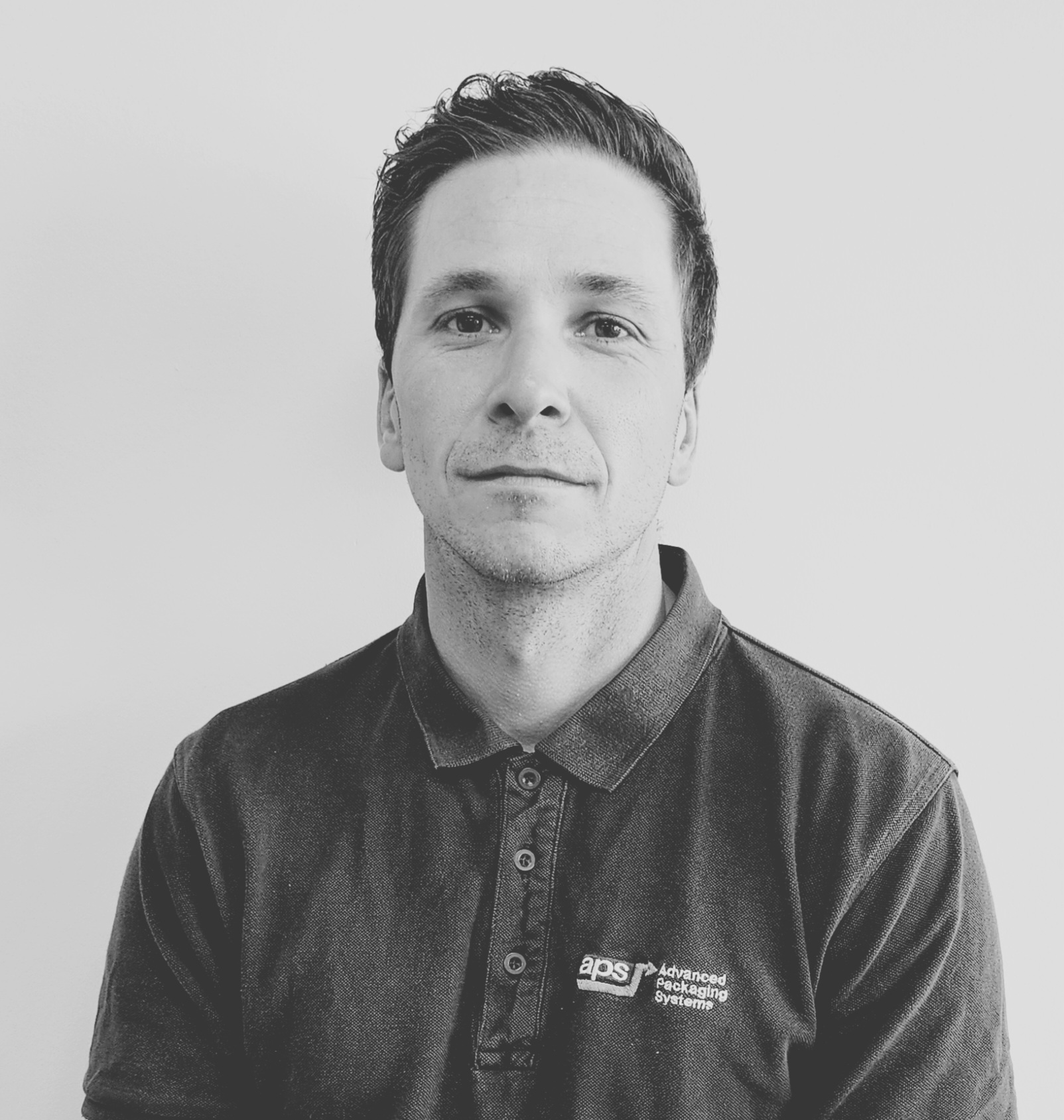 Mike Rushworth - Advanced Packaging Systems Ltd. A man stands against a white background, wearing a dark polo shirt with a logo that reads aps Advanced Packaging Systems. He has short hair and a neutral expression. The photo is in black and white.