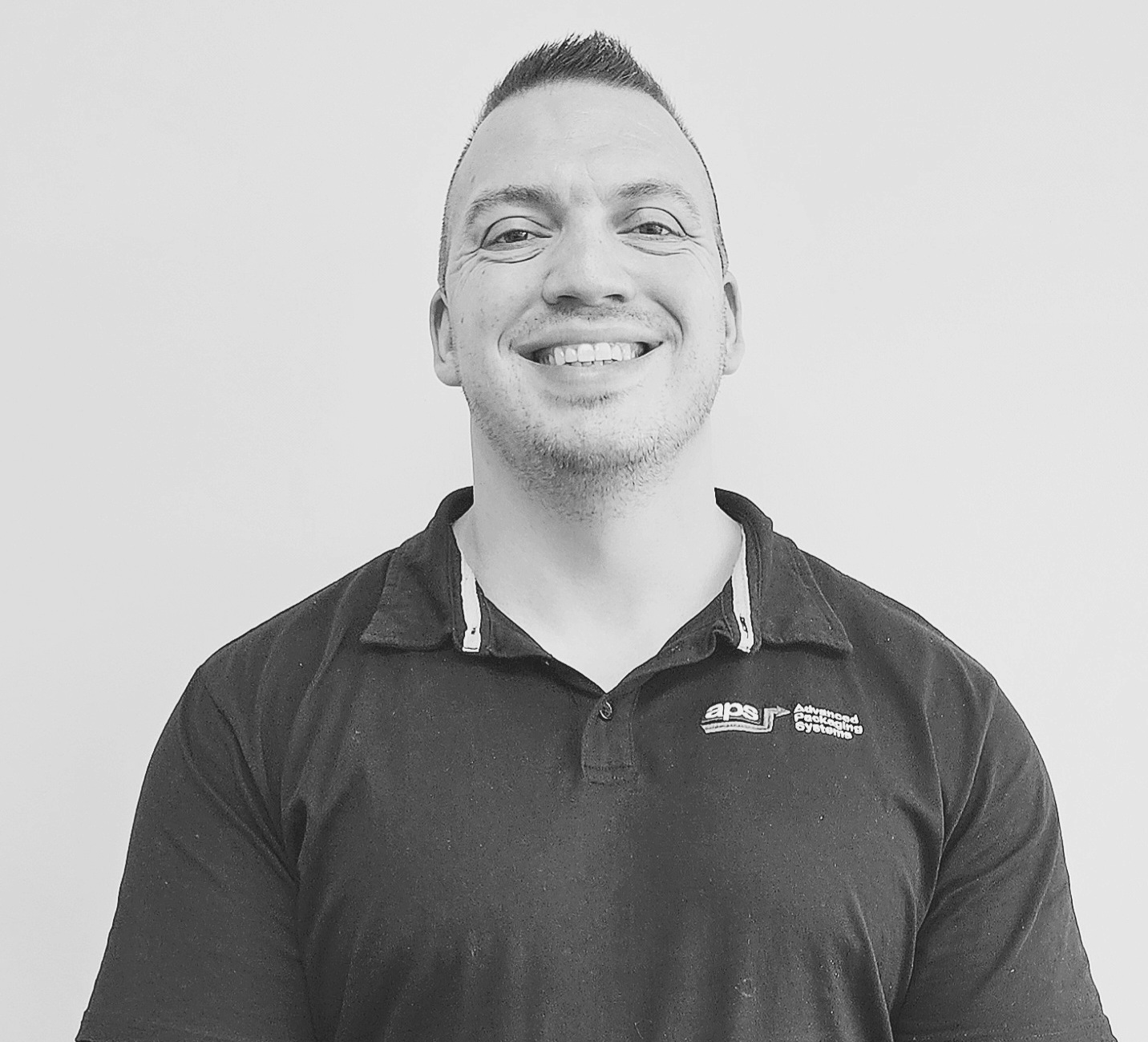 Smiling male person wearing a dark polo shirt with an APS logo on the left side, standing against a plain white background.