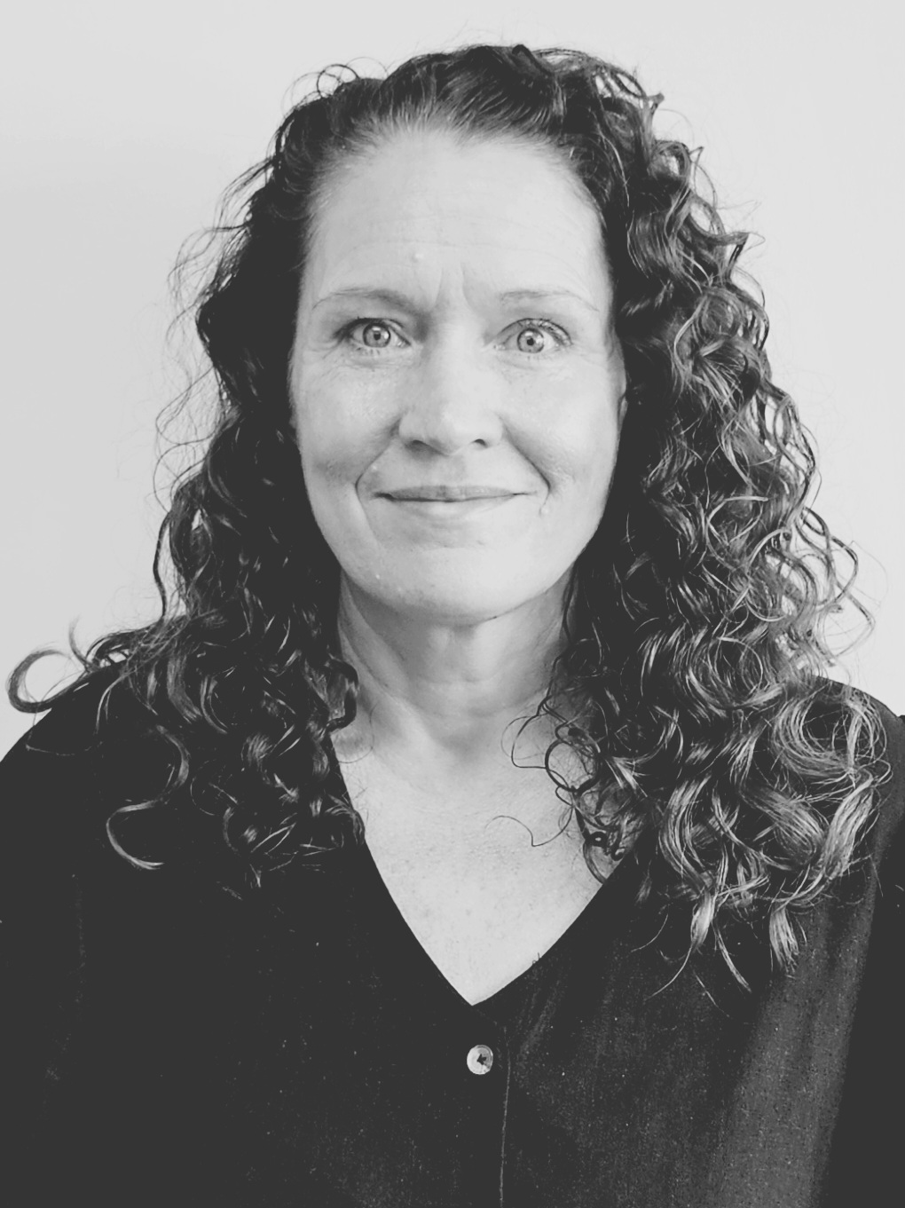 Woman with curly hair and a friendly expression, wearing a dark button-up top, against a plain background in a black and white image.