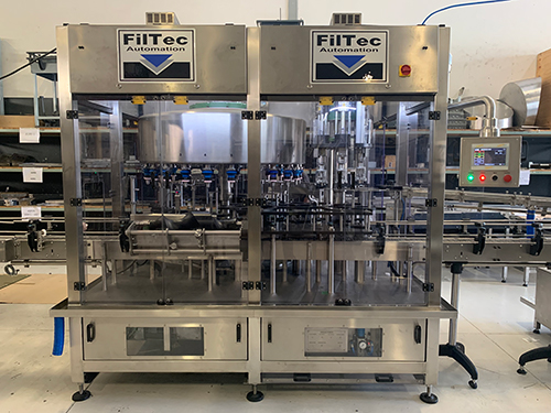 GLF SERIES - GRAVITY LEVEL FILLERS. A stainless steel Filtec automated liquid filling machine with transparent panels, control panel on the right, conveyor belts, and FilTec Automation branding at the top, located in an industrial setting.