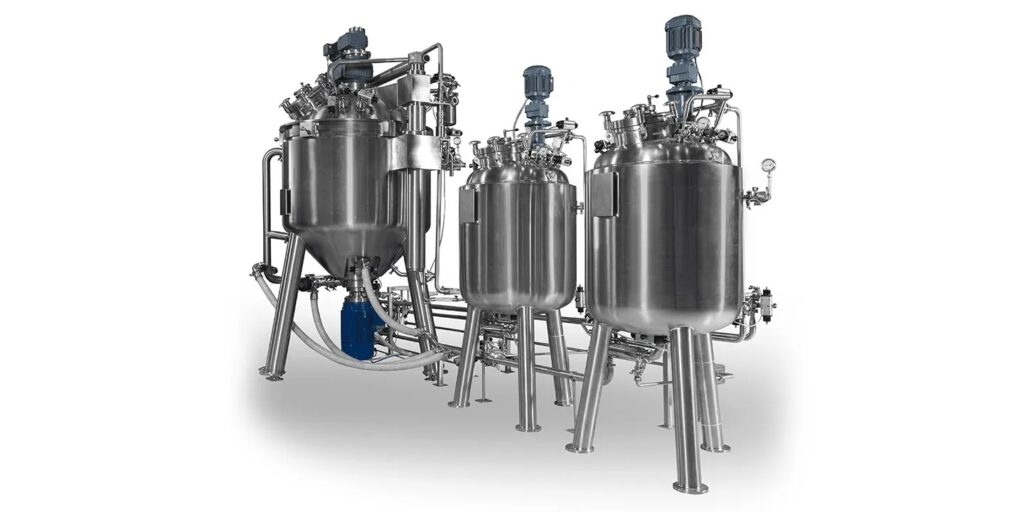 Olsa, tanks and vessels. Three large, stainless steel industrial tanks and vessels with various pipes, valves, and blue electric motors attached, stand on metal legs and are connected as part of a processing system.