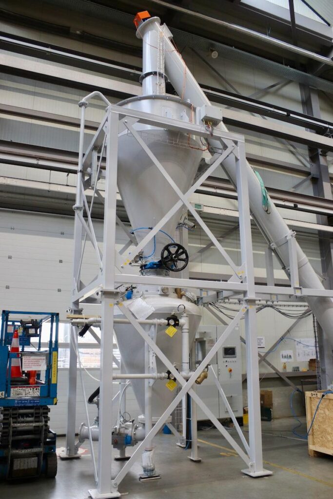 A large industrial cyclone separator by Dinnissen in a warehouse, mounted on a metal frame with pipes connected at the top and bottom, surrounded by equipment and storage boxes.